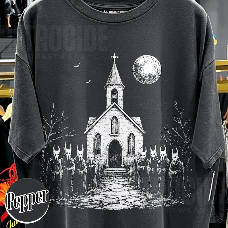 Op de afbeelding: Een antracietgrijs T-shirt met korte mouwen en een witte grafische print van een kerk met een toren, een volle maan en twee vleermuizen. Op de voorgrond staan acht figuren met kappen en gehoornde maskers op een stenen pad.
