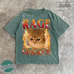 Puede incluir: Una camiseta de manga corta de color verde claro con un gr&aacute;fico de un gato rodeado de llamas naranjas y amarillas. El texto RAGE est&aacute; impreso en rojo sobre el gato, y el texto CONSUMES ME est&aacute; impreso en rojo debajo en una fuente negrita.