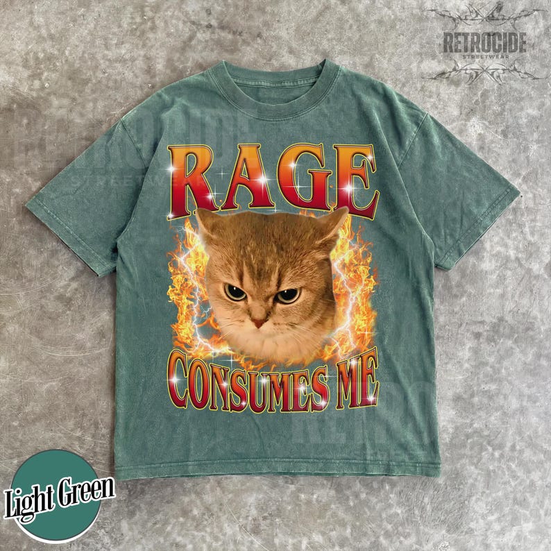 Puede incluir: Una camiseta de manga corta de color verde claro con un gr&aacute;fico de un gato rodeado de llamas naranjas y amarillas. El texto RAGE est&aacute; impreso en rojo sobre el gato, y el texto CONSUMES ME est&aacute; impreso en rojo debajo en una fuente negrita.