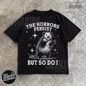 May include: Black t-shirt with a distressed graphic of an opossum and the text "THE HORRORS PERSIST BUT SO DO I." The design includes stars and is printed in white. The shirt has a crew neck and short sleeves.