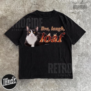 Puede incluir: Una camiseta negra de manga corta con el gr&aacute;fico de un gato blanco y negro tumbado. El texto "live, laugh, loaf" est&aacute; impreso en un patr&oacute;n de llamas de color naranja y amarillo junto al gato. La camiseta tiene cuello redondo y mangas cortas.