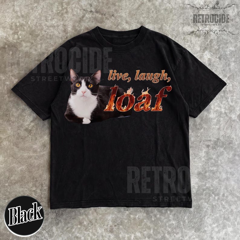Puede incluir: Una camiseta negra de manga corta con el gr&aacute;fico de un gato blanco y negro tumbado. El texto "live, laugh, loaf" est&aacute; impreso en un patr&oacute;n de llamas de color naranja y amarillo junto al gato. La camiseta tiene cuello redondo y mangas cortas.