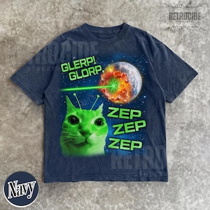 May include: Navy blue t-shirt with a graphic of a green cat with antennae, a planet being zapped by a laser, and the words "GLERP! GLORP." and "ZEP ZEP ZEP." The shirt has a vintage, distressed look.