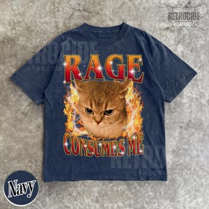 Puede incluir: Una camiseta de manga corta azul marino con un gr&aacute;fico de un gato rodeado de llamas naranjas y amarillas. El texto RAGE est&aacute; impreso en rojo sobre el gato, y el texto CONSUMES ME est&aacute; impreso en rojo debajo, ambos con contornos blancos.