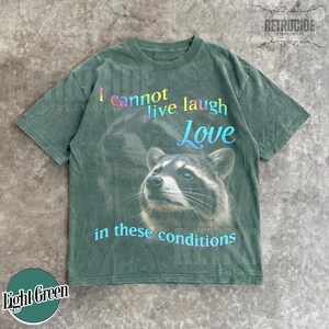 Puede incluir: Una camiseta de manga corta en color verde oliva deste&ntilde;ido con el gr&aacute;fico de un mapache. El texto dice I cannot live laugh Love in these conditions, con la palabra Love en una fuente azul m&aacute;s grande y las otras palabras en degradado.