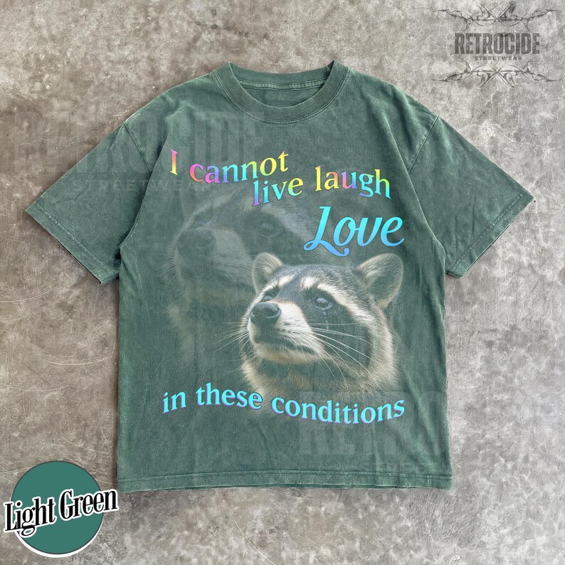Puede incluir: Una camiseta de manga corta en color verde oliva deste&ntilde;ido con el gr&aacute;fico de un mapache. El texto dice I cannot live laugh Love in these conditions, con la palabra Love en una fuente azul m&aacute;s grande y las otras palabras en degradado.