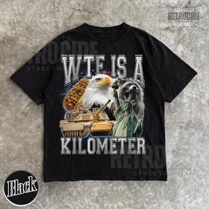 May include: Black t-shirt with a graphic design featuring a hot dog, bald eagle, tank, raccoon, and the Statue of Liberty. The text on the shirt reads "WTF IS A KILOMETER". The shirt has a vintage, distressed look.