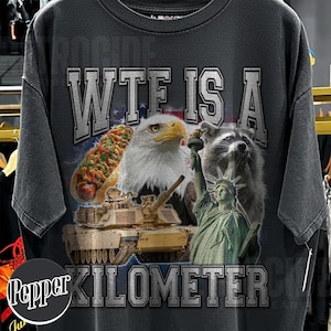 May include: Dark gray t-shirt with a graphic design featuring an eagle, raccoon, hot dog, tank, and the Statue of Liberty. The text "WTE IS A KILOMETER CK" is printed across the top and bottom of the design.