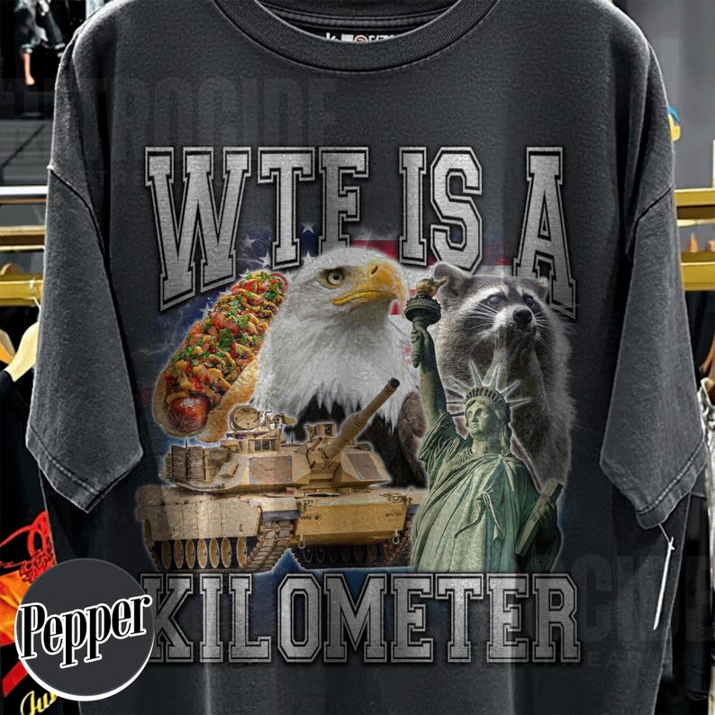 May include: Dark gray t-shirt with a graphic design featuring an eagle, raccoon, hot dog, tank, and the Statue of Liberty. The text "WTE IS A KILOMETER CK" is printed across the top and bottom of the design.