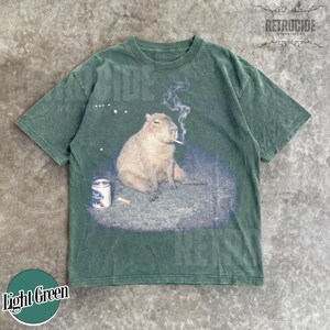 May include: A dark green short-sleeve t-shirt featuring a graphic print of a capybara sitting on the ground with a cigarette in its mouth and a can of Pabst Blue Ribbon beer nearby. The design includes wisps of smoke rising from the cigarette.