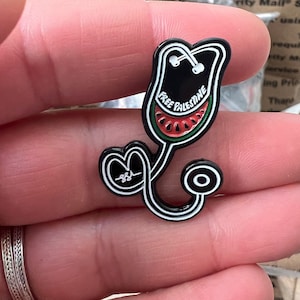 Pode incluir: Pin de esmalte preto em forma de estetoscópio com uma melancia em forma de coração. A melancia tem as palavras "Free Palestine" escritas nela. O estetoscópio tem a forma de um coração com o número "884" escrito dentro.