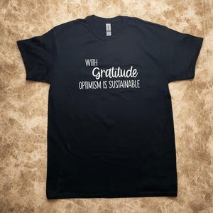 May include: Black t-shirt with the text "WITH Gratitude OPTIMISM IS SUSTAINABLE" in white. The shirt is a crew neck style and appears to be made of a soft material. The shirt is laid flat on a light brown surface.
