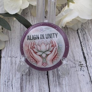 May include: A round, purple button with the text "ALIGN IN UNITY" and a lotus flower design. The design includes hands cradling the flower, with a glittery border. The button is displayed on a clear stand.