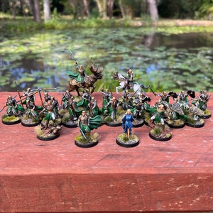 Könnte beinhalten: Eine Sammlung von 20 handbemalten Miniaturfiguren, die Elfen in grüner und blauer Kleidung darstellen, einige reiten auf Pferden und einem Elch, mit Schwertern und Schilden. Die Figuren sind auf einer Holzoberfläche angeordnet.
