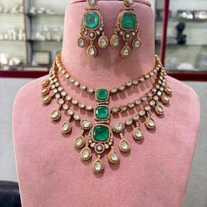 Peut inclure: Un ensemble collier et boucles d'oreilles dorées avec des pierres précieuses vertes taillées en émeraude et des détails clairs en forme de goutte. Le collier comporte plusieurs rangs avec un pendentif central en émeraude. Les boucles d'oreilles reprennent le design.