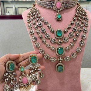 Puede incluir: Un elaborado conjunto de joyas con gargantilla, collar de varias capas y pendientes. Las piezas están adornadas con piedras preciosas transparentes y verde esmeralda, y una piedra rosa. Las joyas se exhiben en un maniquí rosa.