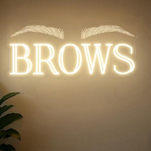 以下が含まれることがあります： 温白色の「BROWS」の文字と、その上にスタイリッシュな眉の形が描かれたネオンサイン。ベージュ色の壁に取り付けられています。受付カウンターと鉢植えの植物が前景に見え、落ち着いた雰囲気を作り出しています。