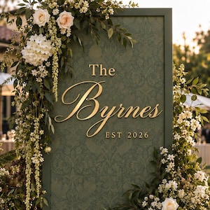 Op de afbeelding: Een hoog, rechthoekig, donkergroen trouwbord met een subtiel damastpatroon en gouden letters met de tekst The Byrnes EST 2026. Het bord is versierd met trapsgewijze arrangementen van witte en lichtroze bloemen en groen gebladerte.