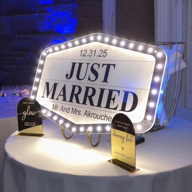 K&ouml;nnte beinhalten: Ein beleuchtetes "Just Married" Schild mit dem Datum 12.31.25. Das Schild ist von Lichtern umgeben und tr&auml;gt die Namen "Mr. And Mrs. Akrouche" unter dem Text. Zwei kleinere Schilder lauten "Let Love Glow" und "A Little Treat For Your Dancing Feet."