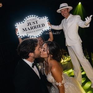 K&ouml;nnte beinhalten: Ein frisch verheiratetes Paar k&uuml;sst sich, mit einem Schild mit der Aufschrift "JUST MARRIED", das &uuml;ber ihnen gehalten wird. Das Schild zeigt das Datum "04.19.2026" und die Namen "JESSICA & JOHNNY". Eine Person in wei&szlig;em Anzug und Hut l&auml;chelt im Hintergrund.