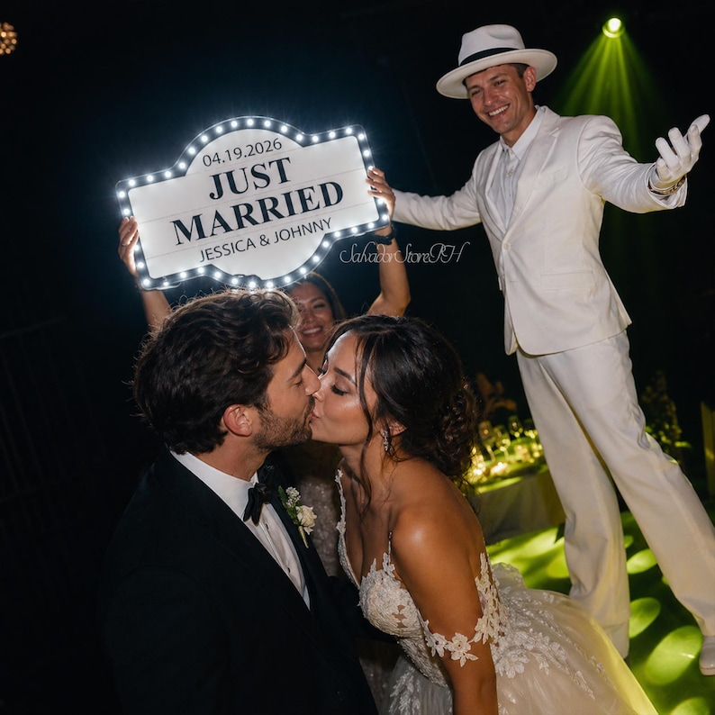 K&ouml;nnte beinhalten: Ein frisch verheiratetes Paar k&uuml;sst sich, mit einem Schild mit der Aufschrift "JUST MARRIED", das &uuml;ber ihnen gehalten wird. Das Schild zeigt das Datum "04.19.2026" und die Namen "JESSICA & JOHNNY". Eine Person in wei&szlig;em Anzug und Hut l&auml;chelt im Hintergrund.