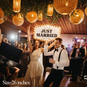 K&ouml;nnte beinhalten: Eine Hochzeitsfeier mit einem Paar, das ein Schild mit der Aufschrift "JUST MARRIED" h&auml;lt. Das Schild ist 66 cm breit und hat einen beleuchteten Rand. Das Paar ist von G&auml;sten und dekorativen H&auml;ngelampen umgeben.