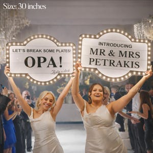 K&ouml;nnte beinhalten: Zwei beleuchtete Schilder, jeweils etwa 76 cm gro&szlig;, werden auf einer Hochzeit hochgehalten. Auf einem Schild steht "LET'S BREAK SOME PLATES! OPA!" und auf dem anderen "INTRODUCING MR & MRS PETRAKIS". Die Schilder haben einen Vintage-Marquee-Stil mit Gl&uuml;hbirnen.