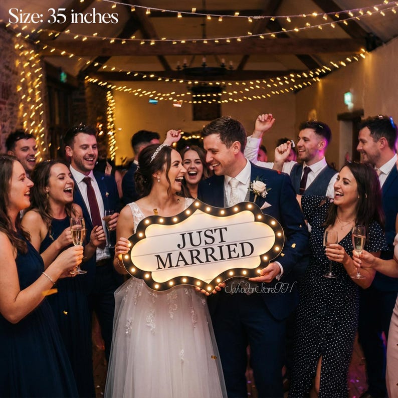 K&ouml;nnte beinhalten: Ein frisch verm&auml;hltes Paar h&auml;lt ein 89 cm gro&szlig;es Schild mit der Aufschrift &bdquo;JUST MARRIED&ldquo; und beleuchteten Lichtern. Das Schild hat einen dekorativen Rand und wird von l&auml;chelnden Hochzeitsg&auml;sten umgeben. Das Schild ist wei&szlig;.