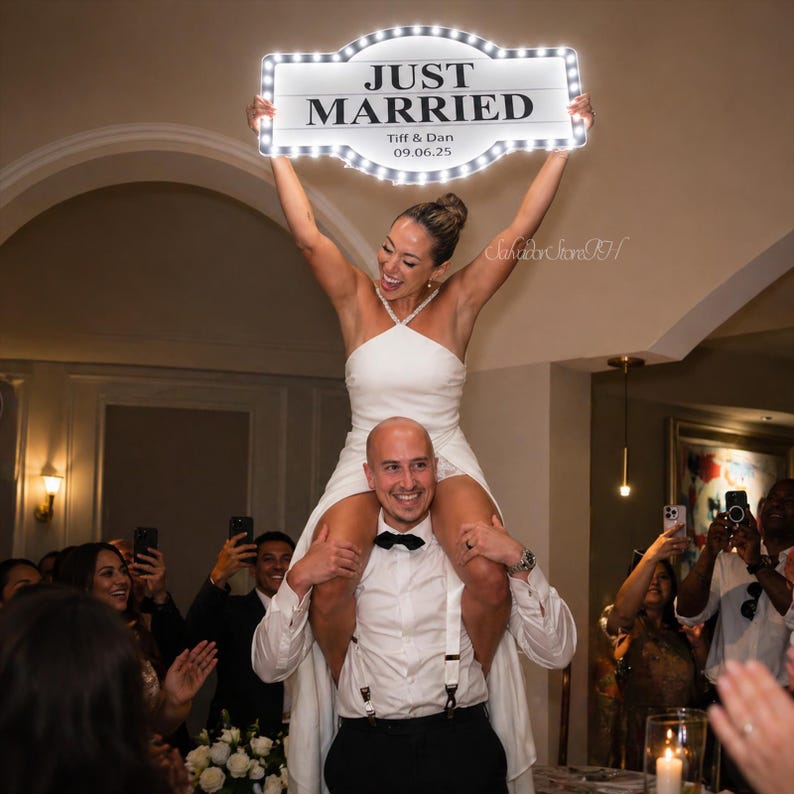 May include: A newly married couple celebrates, with the bride perched on the groom's shoulders. She holds a light-up sign saying "JUST MARRIED," with the names "Tiff & Dan" and the date "09.06.25." Guests are visible in the background.