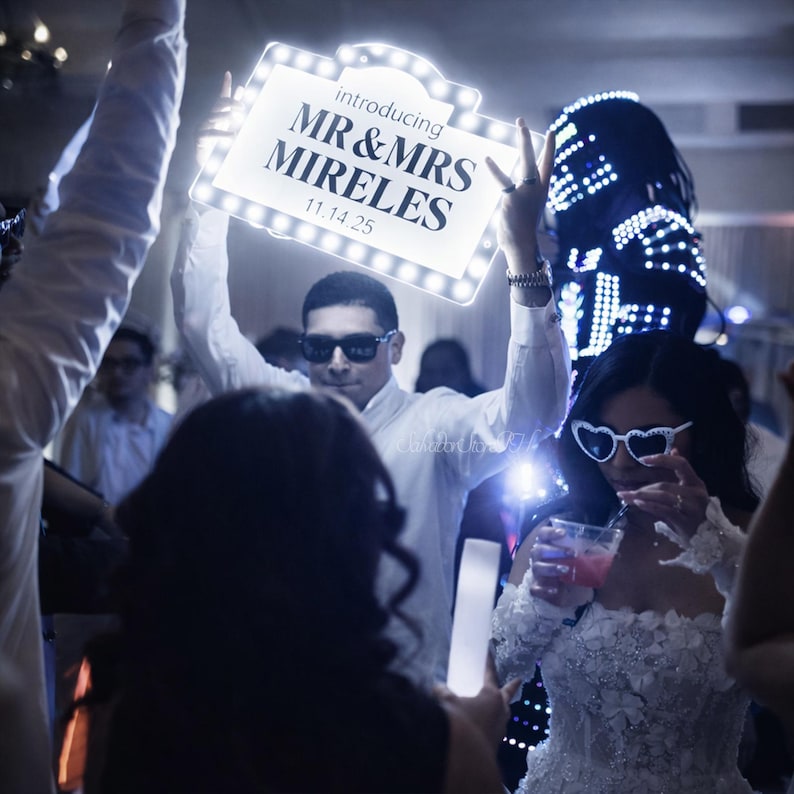 K&ouml;nnte beinhalten: Ein beleuchtetes Schild mit dem Text "introducing MR & MRS MIRELES 11.14.25" wird auf einer Hochzeitsfeier hochgehalten. Das Schild ist von hellen Lichtern umgeben. Eine Person mit Sonnenbrille h&auml;lt das Schild hoch. Eine Frau in einem wei&szlig;en Kleid und herzf&ouml;rmiger Sonnenbrille trinkt ein Getr&auml;nk.