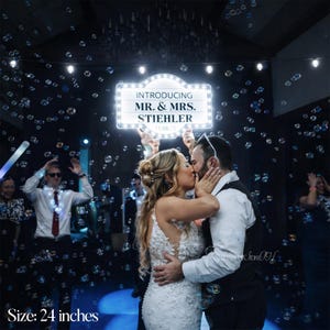 K&ouml;nnte beinhalten: Eine Hochzeitsfeier mit einem sich k&uuml;ssenden Paar unter einem Schild mit der Aufschrift "INTRODUCING MR. & MRS. STIEHLER". Das Schild ist 61 cm gro&szlig; und von Lichtern umgeben. Blasen schweben in der Luft.