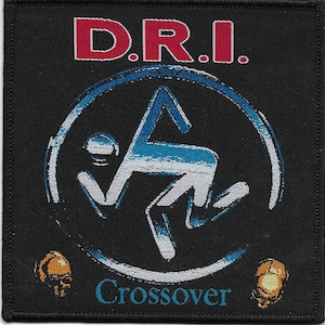 May include: A square black embroidered patch featuring the red text D.R.I. at the top. The center displays a blue and white stylized figure inside a circle, with the blue text Crossover below. Two small orange skull icons are at the bottom corners.