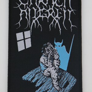 May include: Black and white embroidered patch with the band name "Carach Angren" in a gothic font. The patch features a blue silhouette of a person sitting on a wooden floor with a blue devil-like figure behind them.