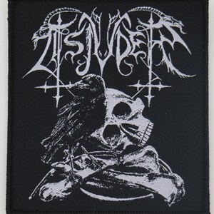 May include: Black and white embroidered patch with the band name "Tsjuder" in a gothic font. The patch features a skull with a crow perched on top, and a stylized design of a cross with wings.