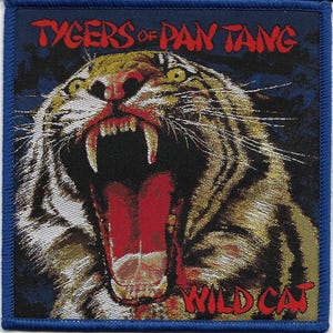 May include: A square patch with a roaring tiger design. The patch features the text "TYGERS OF PAN TANG" in red at the top and "WILD CAT" at the bottom. The tiger is depicted with a wide-open mouth, showcasing its teeth. The patch has a blue border.