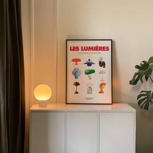 May include: A white table lamp with a round, glowing, yellow-orange shade. A framed poster with the text "LES LUMI&Egrave;RES" and various colorful lamp designs is on a white cabinet. The room has a neutral color palette.