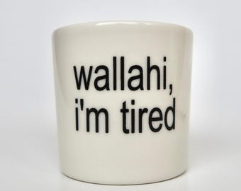 Op de afbeelding: Een witte keramische mok met de zwarte gedrukte tekst wallahi, i'm tired op de voorzijde. De mok heeft een cilindrische vorm met een gladde afwerking en een eenvoudig, strak ontwerp, waarbij de tekst duidelijk in een schreefloos lettertype staat.