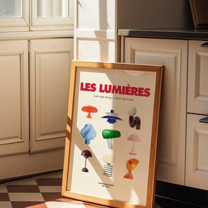 May include: A framed art print featuring colorful illustrations of various lamps. The text "LES LUMI&Egrave;RES" is displayed in red, with the tagline "I call it light design, my bank calls it debt." The print is in a light wood frame.