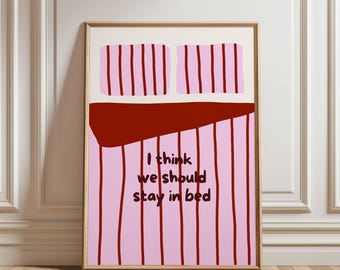 Op de afbeelding: Een ingelijste kunstprint met een gestileerde bedillustratie. Het bed heeft roze gestreepte beddengoed en twee roze kussens. De tekst "I think we should stay in bed" is in het zwart geschreven. De lijst is lichtbruin.
