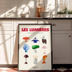 May include: A framed poster featuring a collection of colorful, stylized lamps. The poster's title, "LES LUMI&Egrave;RES," is in red, with the text "I call it light design, my bank calls it debt." The bottom of the poster reads "LEVKALADE STUDIO EST. 2019."