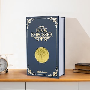May include: A navy blue book embosser with gold lettering and a gold seal. The book is standing upright on a wooden surface. The text on the book reads "BOOK EMBOSSER" and "Edit By CasaFit".