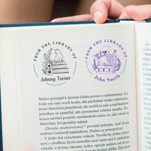 May include: Two custom library stamps are shown on a book page. One stamp features a cat on books with flowers, the other a skeleton reading. Both stamps are circular with the text "FROM THE LIBRARY OF" and a name.