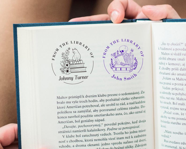 May include: Two custom library stamps are shown on a book page. One stamp features a cat on books with flowers, the other a skeleton reading. Both stamps are circular with the text "FROM THE LIBRARY OF" and a name.