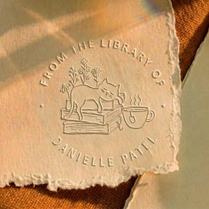 May include: Cream-colored paper with a debossed design featuring a cat resting on books, a teacup, and flowers. The text "FROM THE LIBRARY OF DANIELLE PATEL" is embossed around the design. The paper has a deckled edge.