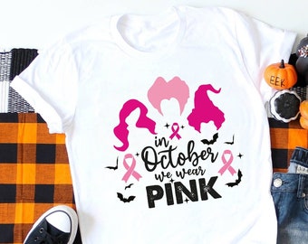 Op de afbeelding: Wit T-shirt met een roze lint en de tekst "in October we wear PINK" en een afbeelding van drie heksen met roze haar en hoeden. Het shirt ligt op een geruite deken met een paar zwarte sneakers en een blauwe jeans.
