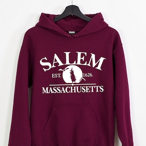 May include: A maroon hoodie with a white graphic print that says "SALEM EST. 1626. MASSACHUSETTS" with a silhouette of a witch flying on a broomstick in front of a full moon.
