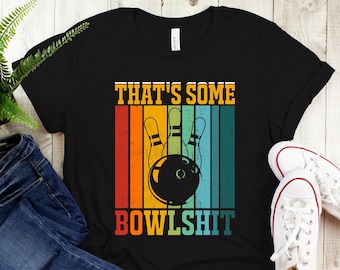 May include: Black t-shirt with the text "THAT'S SOME BOWLSHT" in a retro design featuring bowling pins and a bowling ball. The shirt is paired with blue jeans and white sneakers.