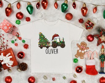 May include: White t-shirt featuring a Christmas-themed graphic. The design includes a green tractor pulling a trailer with a Christmas tree, a Santa hat, and the name "OLIVER" in black. The shirt is surrounded by Christmas decorations.