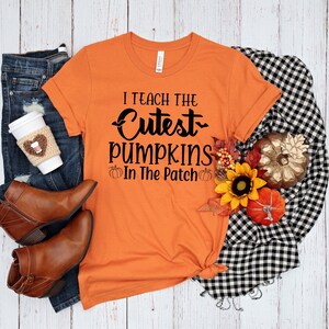 May include: Orange t-shirt with black text that reads "I teach the cutest pumpkins in the patch". The shirt is on a white wooden surface with a pair of brown boots, blue jeans, a coffee cup, and a plaid blanket.