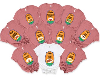 May include: Ten pink t-shirts with different sauce-themed designs. Each shirt features a label with a different sauce name and a small illustration of the sauce. The shirts are arranged in a circle with a white t-shirt in the center. The white t-shirt has a steak sauce design.